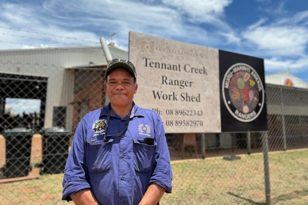 An Aboriginal man wearing a cap and blue work shirt looks at the camera in front a Tennant Creek Ranger Workshed sign.