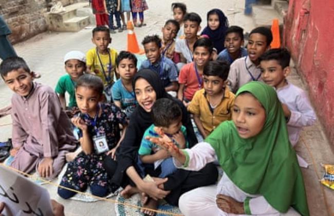 A score of smiling young children sit on the ground in an alleyway as someone off camera reads to them 