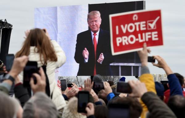 People are seen from the back in the crowd and look toward a screen showing an older man in a black suit and red tie. One person holds a sign that says 'I vote pro-life.'