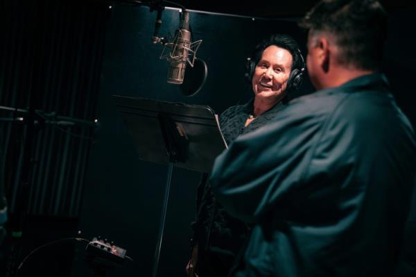 Wayne Newton in a black jacket and headpho<em></em>nes recording vocals for the song 'This Is America' next to a microphone