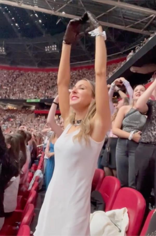 Nikki Glaser in a white dress, raising her arms in a crowd at a Taylor Swift concert