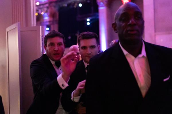 Alex Bruesewitz being escorted backstage after collapsing on stage at the New York Young Republican Club's Annual Gala, surrounded by men in suits